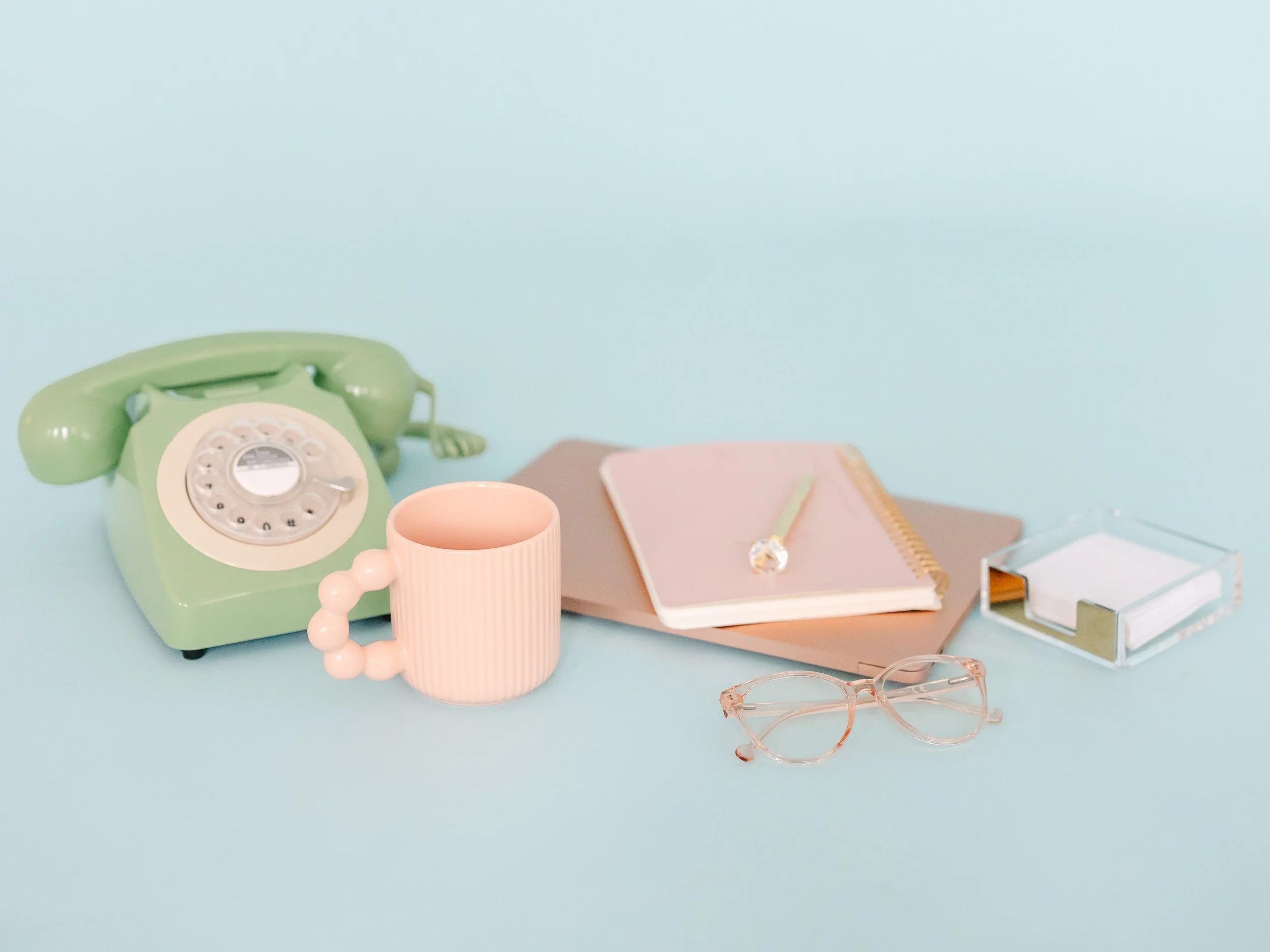 creative virtual assistant desk with pink laptop, green dial phone, pink mug, notebook and glasses
